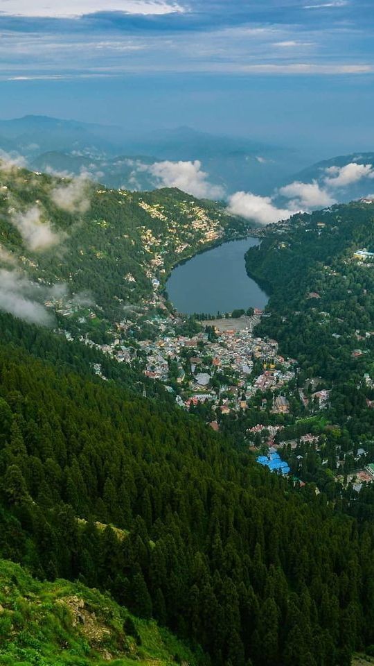 A mesmerizing aerial view of a serene hill town nestled around a tranquil lake, surrounded by lush green forests and misty mountains that capture the timeless charm of Uttarakhand’s hill stations.